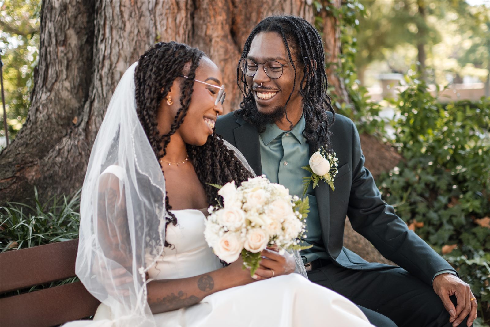Bride and groom laughing together on a park bench under the trees