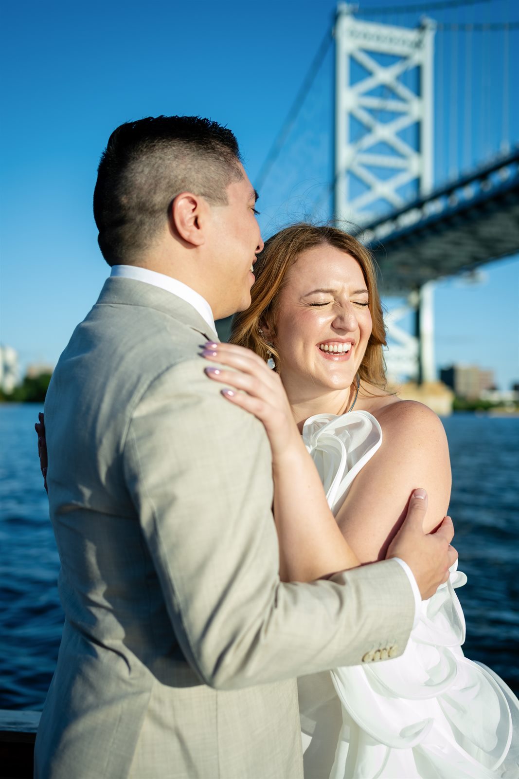 Bride laughing while embracing her groom with the Benjamin Franklin Bridge behind them