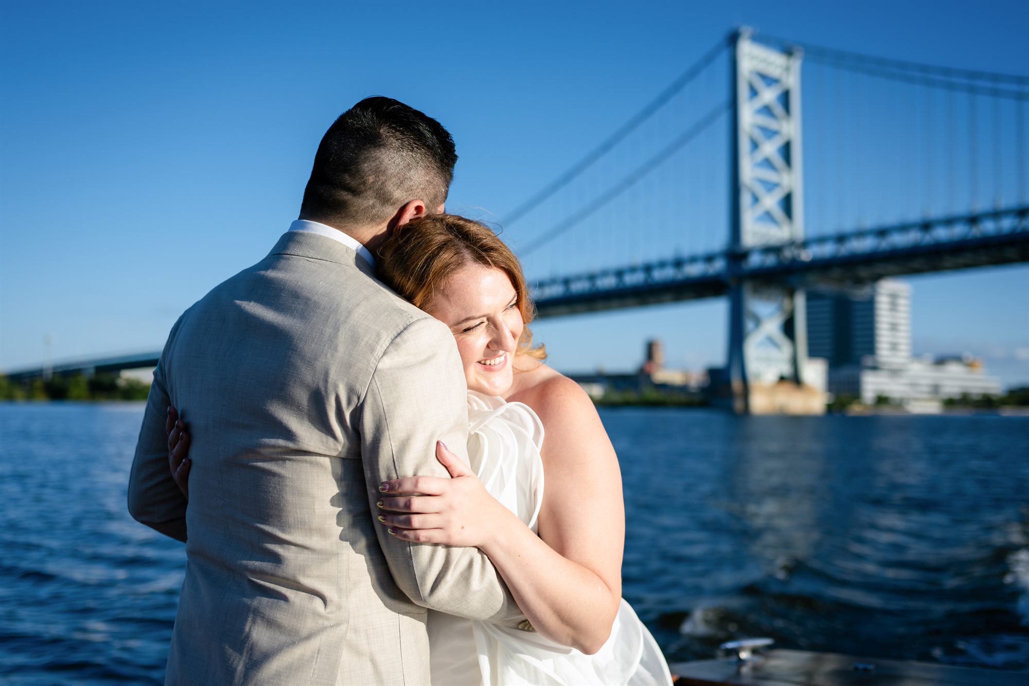 Bride and groom embracing on the Delaware River waterfront with the full span of the Benjamin Franklin Bridge behind them