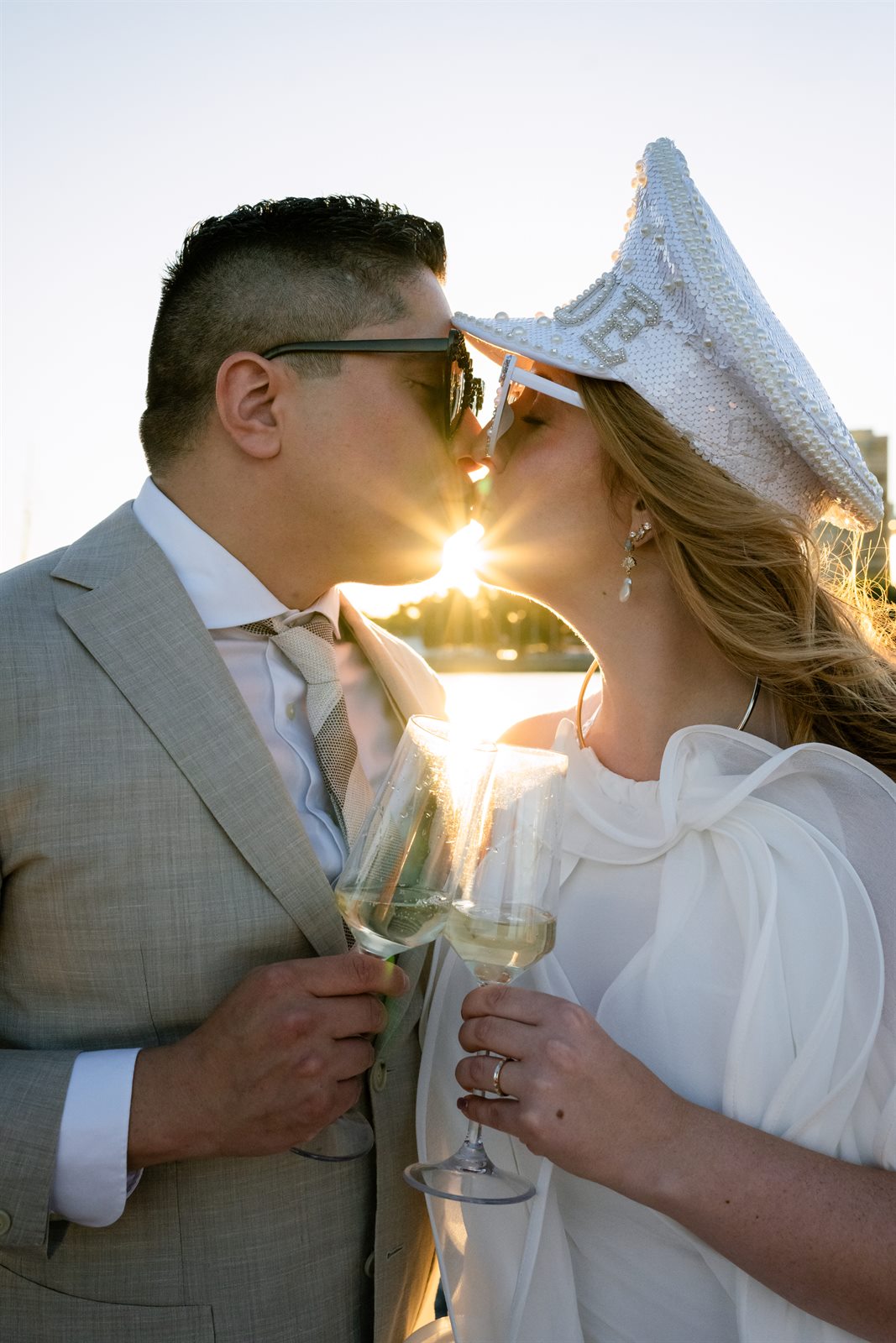 Bride and groom kissing over a champagne toast at sunset, sun flare between them