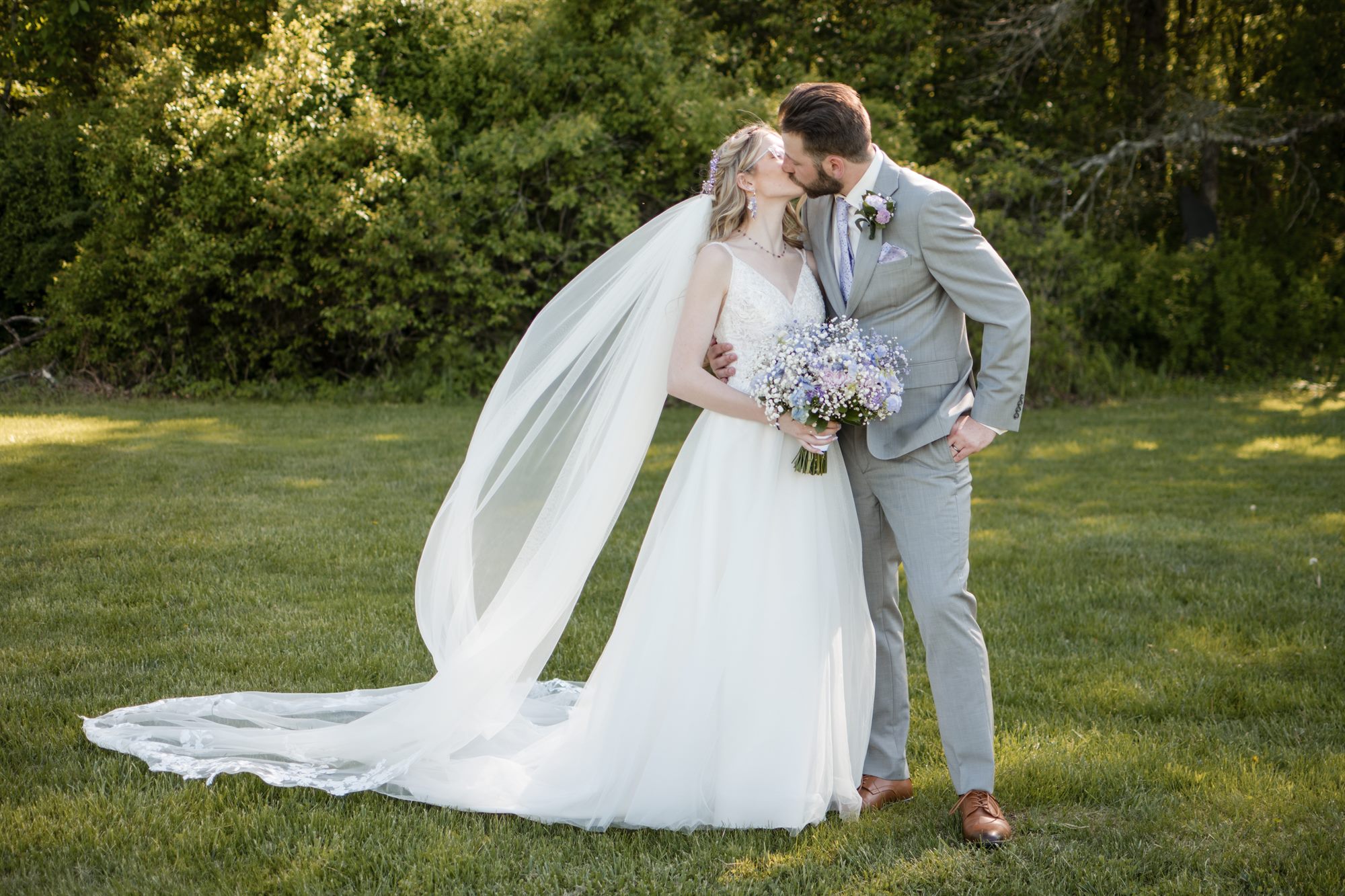 Bride and groom kissing in a field while her veil catches the wind