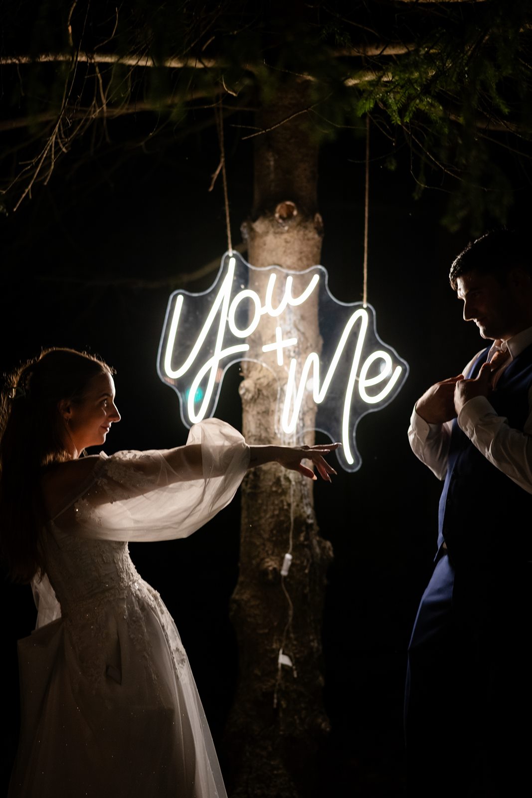 Bride reaching toward a glowing You + Me neon sign in the trees while her groom straightens his vest at night